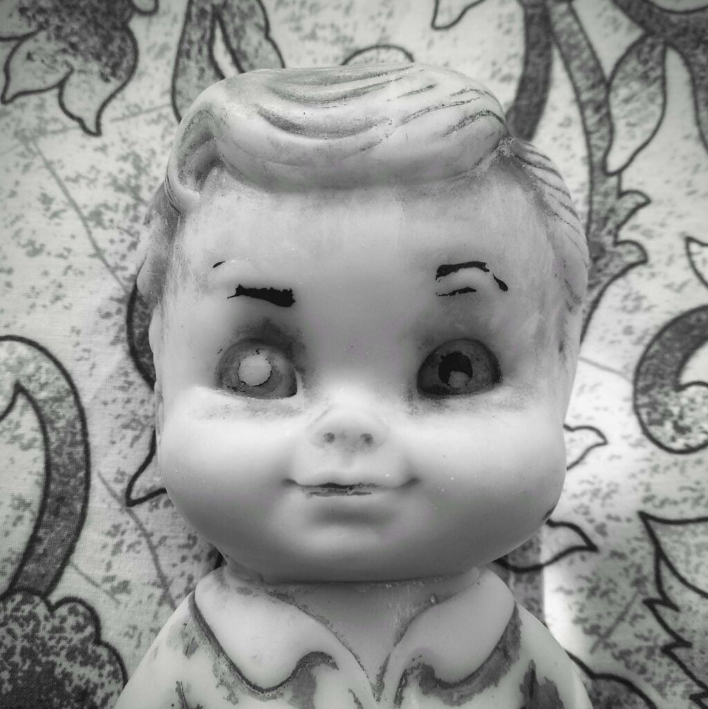 Black-and-white close-up photo of a vintage bisque baby doll's face with painted eyebrows and glass eyes, one eye chipped, against a floral fabric background.