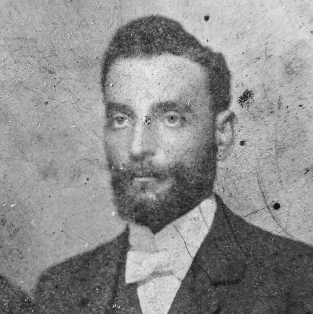 Weathered black-and-white portrait photo of a man with a full beard in a suit and bow tie, damaged by scratches and stains.