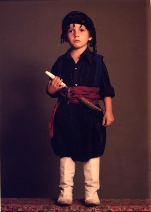 Studio photo of a young child dressed in traditional Greek costume, a dark shirt with a red sash and prop knife and white boots, standing on a patterned rug.